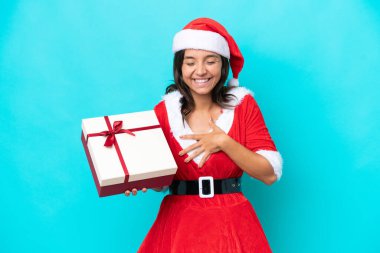 Anne Noel gibi giyinmiş genç İspanyol kadın elinde mavi arka planda izole edilmiş bir hediyeyle gülümsüyor.