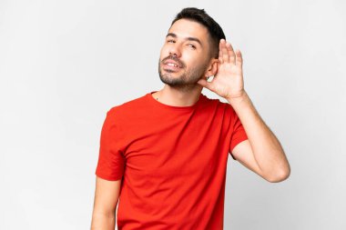 İzole edilmiş beyaz arka planda genç, yakışıklı beyaz bir adam elini kulağına koyarak bir şeyler dinliyor.