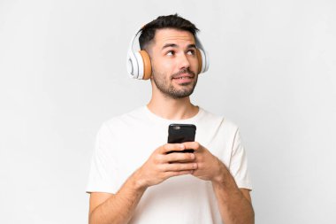 Young handsome caucasian man over isolated white background listening music with a mobile and thinking