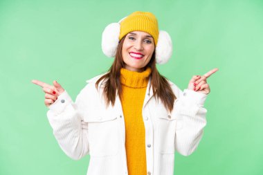 Middle age woman wearing winter muffs over isolated chroma key background pointing finger to the laterals and happy