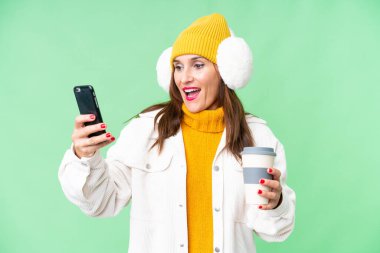 Orta yaşlı bir kadın, izole edilmiş krom arka planda kış maskesi takıyor. Elinde kahve ve cep telefonu var.