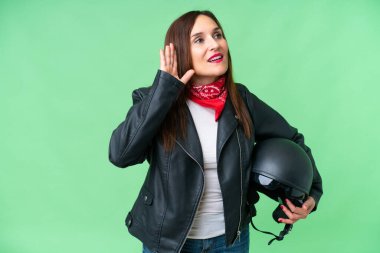 Middle age caucasian woman with a motorcycle helmet over isolated chroma key background listening to something by putting hand on the ear