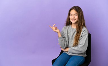Little caucasian girl sitting on a chair isolated on purple background happy and pointing up