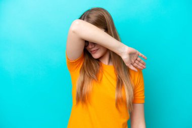 Mavi arka planda izole edilmiş genç beyaz kadın gözlerini elleriyle kapatıyor.