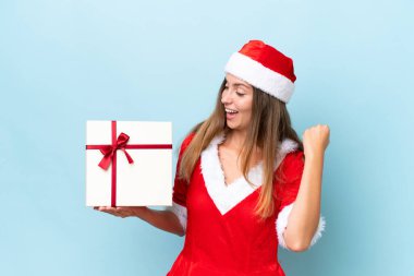 Young caucasian woman dressed as mama noel holding gift isolated on blue background celebrating a victory