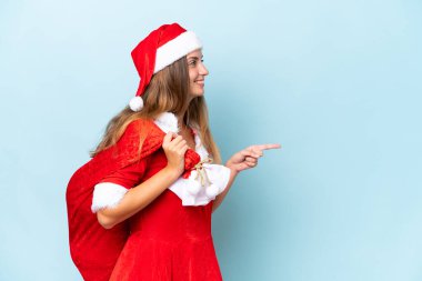 Anne Noel gibi giyinmiş, mavi arka planda izole edilmiş Noel çuvalını tutan beyaz bir kadın bir ürünü sunmak için kenarı işaret ediyor.