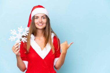 Anne Noel gibi giyinmiş, elinde kar taneleri olan beyaz bir kadın mavi arka planda izole edilmiş bir şekilde bir ürünü sunmak için kenarı işaret ediyor.