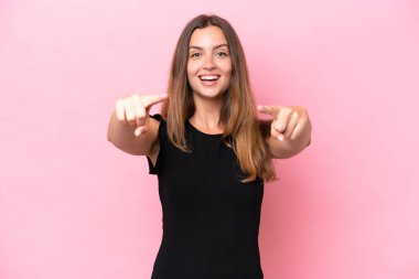 Pembe arka planda izole edilmiş genç beyaz kadın şaşırmış ve ön tarafı işaret ediyor.