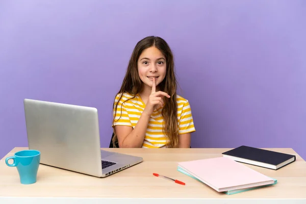 Little student girl in a workplace with a laptop isolated on purple background showing a sign of silence gesture putting finger in mouth