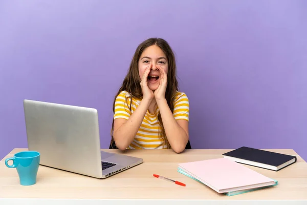 Little student girl in a workplace with a laptop isolated on purple background shouting and announcing something