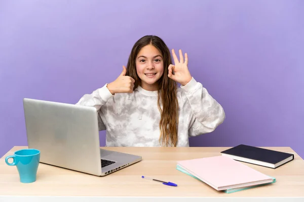 Little student girl in a workplace with a laptop isolated on purple background showing ok sign and thumb up gesture