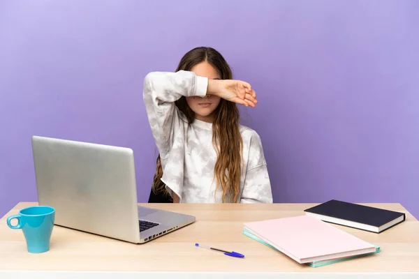 Little student girl in a workplace with a laptop isolated on purple background covering eyes by hands