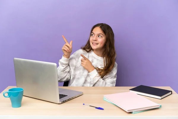 Little student girl in a workplace with a laptop isolated on purple background pointing with the index finger a great idea