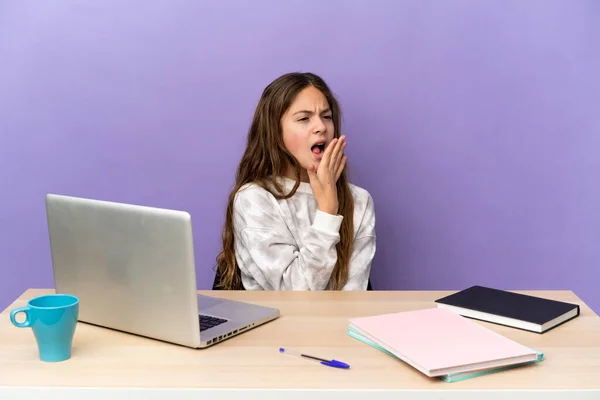 Little student girl in a workplace with a laptop isolated on purple background yawning and covering wide open mouth with hand