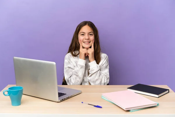 Little student girl in a workplace with a laptop isolated on purple background smiling with a happy and pleasant expression