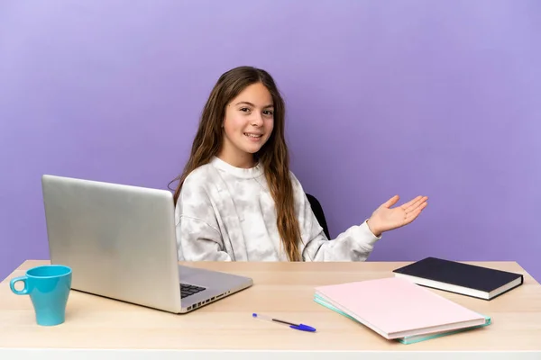 Little student girl in a workplace with a laptop isolated on purple background extending hands to the side for inviting to come