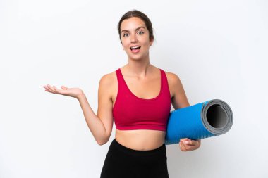 Beyaz arka planda, şok olmuş bir yüz ifadesiyle izole edilmiş bir minderi tutarken yoga derslerine giden beyaz bir kadın.