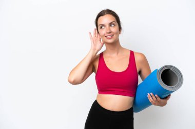 Young sport caucasian woman going to yoga classes while holding a mat isolated on white background listening to something by putting hand on the ear