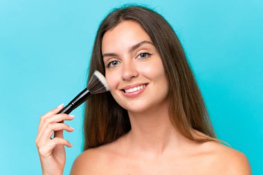 Mavi arka planda izole edilmiş beyaz bir kadın elinde makyaj fırçası ve mutlu bir ifade var.