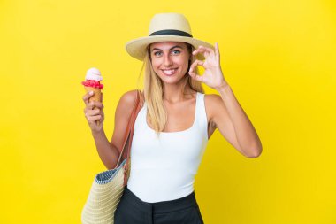 Blonde Uruguayan girl in summertime holding ice cream isolated on yellow background showing ok sign with fingers