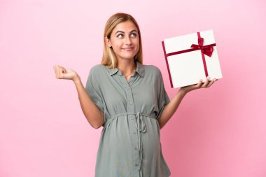 Genç Uruguaylı kadın mavi arka planda izole edilmiş hamile ve elinde şüpheleri varken bir hediye tutuyor.