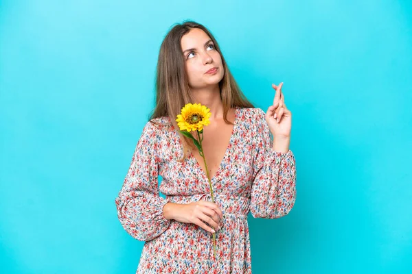 Young caucasian woman holding sunflower isolated on blue background with fingers crossing and wishing the best