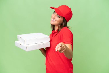 Pizza delivery caucasian woman with work uniform picking up pizza boxes isolated on green chroma background pointing front with happy expression