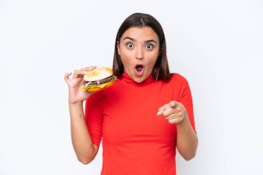 Beyaz arka planda izole edilmiş hamburger tutan beyaz bir kadın şaşırmış ve ön tarafı işaret ediyor.