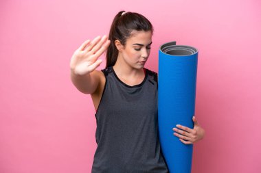 Young Brazilian sport woman going to yoga classes while holding a mat isolated on pink background making stop gesture and disappointed