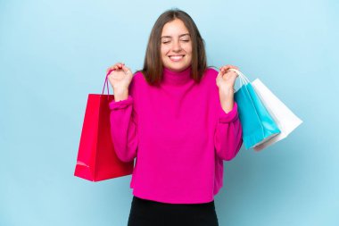 Genç ve güzel bir kadın mavi arka planda izole edilmiş alışveriş torbaları tutuyor ve gülümsüyor.
