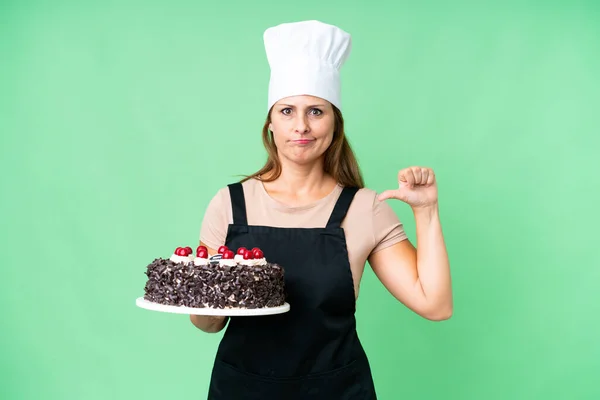 Middle age pastry chef woman holding a big cake over isolated background showing thumb down with negative expression