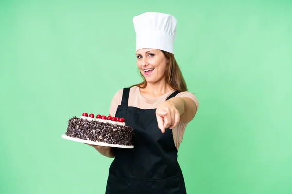 Middle age pastry chef woman holding a big cake over isolated background pointing front with happy expression
