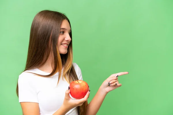 Teenager caucasian girl holding an apple over isolated background pointing to the side to present a product