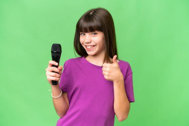 Küçük şarkıcı kız izole edilmiş bir arka planda mikrofonu kaldırıyor. Başparmakları havada çünkü iyi bir şey oldu.