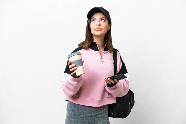 Young sport woman with sport bag over isolated white background holding coffee to take away and a mobile while thinking something