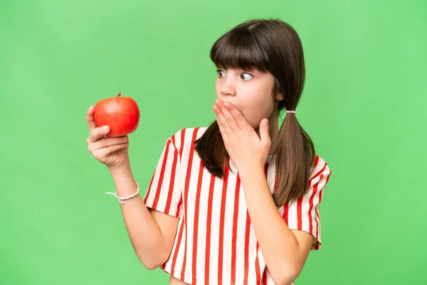 Little caucasian girl holding an apple over isolated background with surprise and shocked facial expression