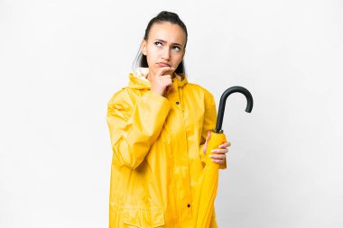 Yağmura dayanıklı ceketi ve şemsiyesi olan genç bir Arap kadını izole edilmiş beyaz arka planda kuşkuları var.