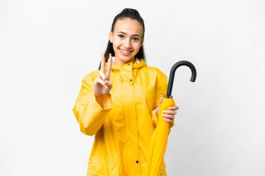 Yağmura dayanıklı ceketli ve şemsiyeli genç Arap kadın izole edilmiş beyaz arka planda gülümsüyor ve zafer işareti gösteriyor.