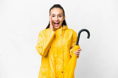 Yağmura dayanıklı ceketi ve şemsiyesi olan genç bir Arap kadını, yalıtılmış beyaz arka planda, şaşırmış ve şok olmuş bir yüz ifadesi var.