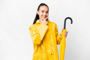 Yağmura dayanıklı ceketi ve şemsiyesi olan genç bir Arap kadını izole edilmiş beyaz arka planda mutlu ve gülümsüyor.