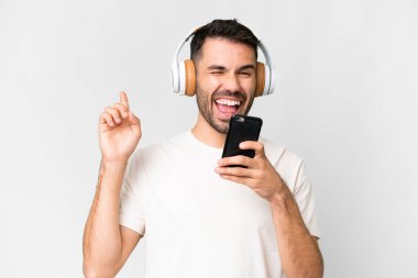 İzole edilmiş beyaz arka planda genç, yakışıklı beyaz bir adam cep telefonuyla müzik dinliyor ve şarkı söylüyor.