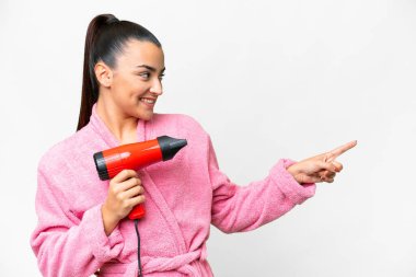 Bornozlu genç bir kadın, elinde saç kurutma makinesiyle izole edilmiş beyaz arka planda bir ürün sunmak için kenarı işaret ediyor.