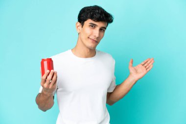 Arjantinli genç adam mavi arka planda izole edilmiş bir içecek tutuyor. Gelmesi için ellerini yana uzatıyor.