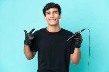 Dövme sanatçısı Arjantinli adam mavi arka planda izole edilmiş bir şekilde baş parmak hareketi yapıyor.