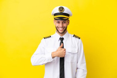 Kafkasyalı uçak pilotu sarı arka planda izole edilmiş bir şekilde baş parmak hareketi yapıyor.