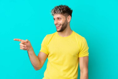 Mavi arka planda izole edilmiş genç, yakışıklı, beyaz bir adam parmağını yana uzatıp bir ürün sunuyor.