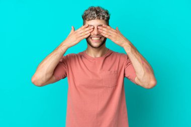 Genç yakışıklı beyaz adam mavi arka planda izole edilmiş gözleri elleriyle kaplıyor ve gülümsüyor.