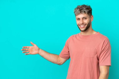 Mavi arka planda izole edilmiş yakışıklı beyaz adam, davet edilmek için ellerini yana uzatıyor.