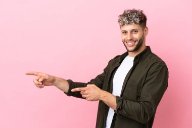 Pembe arka planda izole edilmiş genç, yakışıklı beyaz bir adam parmağını yana uzattı ve bir ürün sundu.