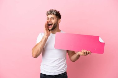 Pembe arka planda izole edilmiş yakışıklı beyaz bir adam boş bir levha tutuyor ve bağırıyor.
